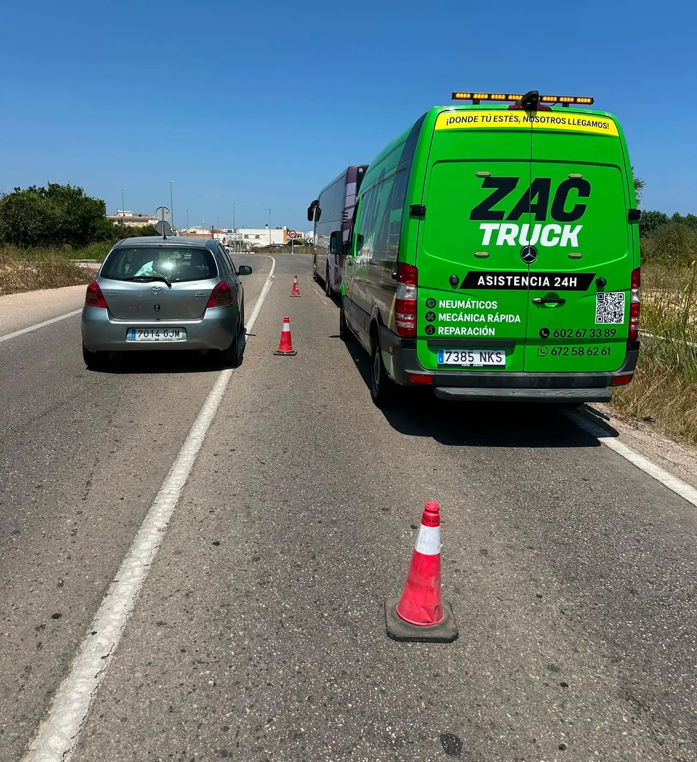 Furgoneta taller de ZAC TRUCK señalizada con conos atendiendo a un coche con una rueda pinchada en el arcén, junto a un autobús